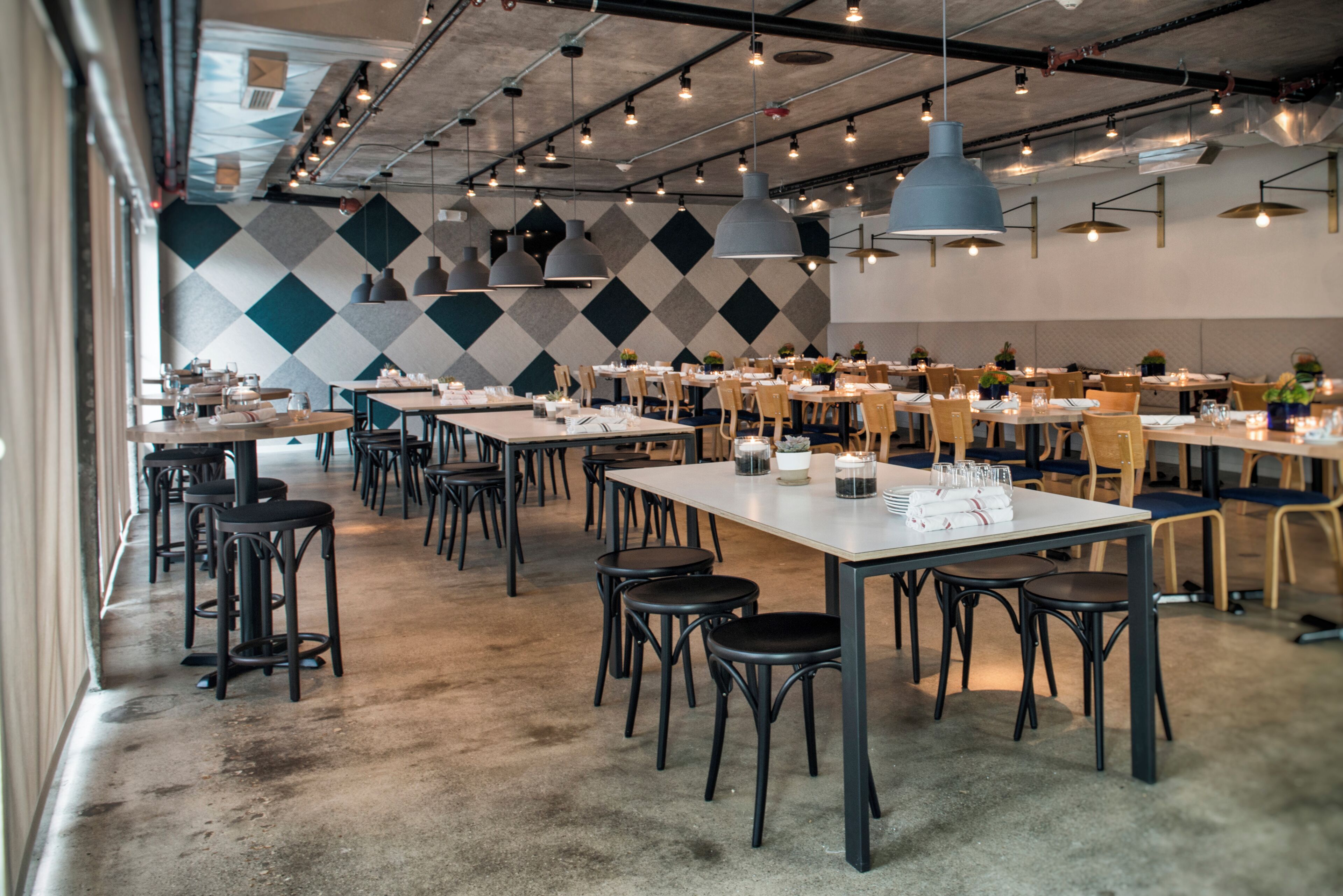 A spacious restaurant with high ceilings, pendant lighting, and a patterned wall. Tables are neatly arranged with chairs, and the floor is concrete.