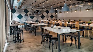 A spacious restaurant with high ceilings, pendant lighting, and a patterned wall. Tables are neatly arranged with chairs, and the floor is concrete.