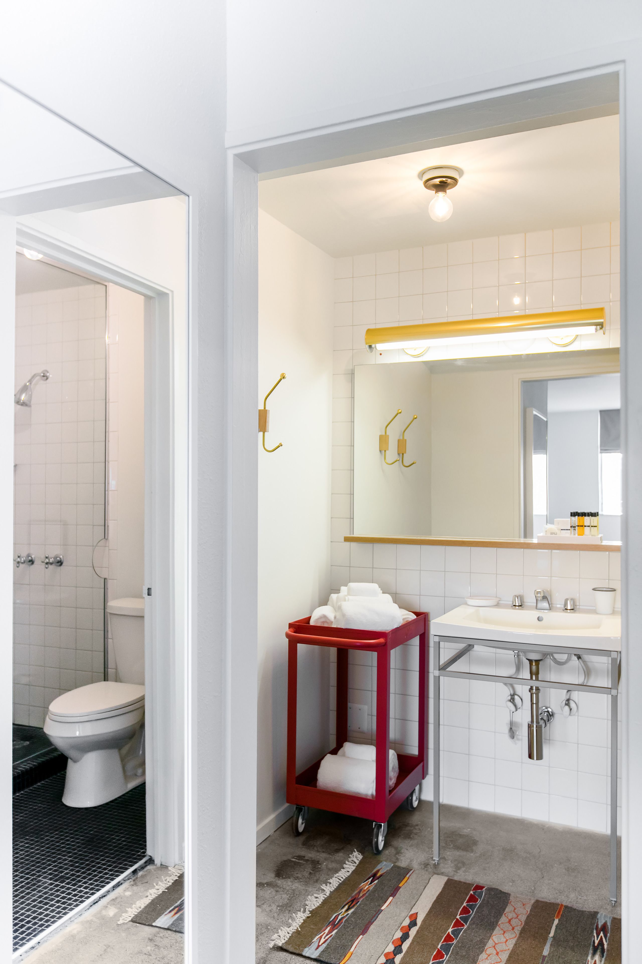 Une salle de bain avec des toilettes blanches, un chariot rouge contenant des serviettes, un lavabo et un miroir.