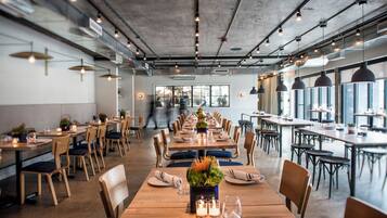 Um restaurante espaçoso com longas mesas de madeira, diversos arranjos de assentos e um design moderno de teto industrial.