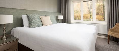 A neatly made bed with a white comforter and patterned pillows, a bedside table with a lamp, a chair, and a window with curtains.