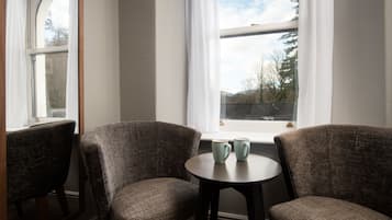 Een kamer met twee fauteuils en een tafeltje met twee kopjes, nabij een raam met uitzicht op bomen en bergen.