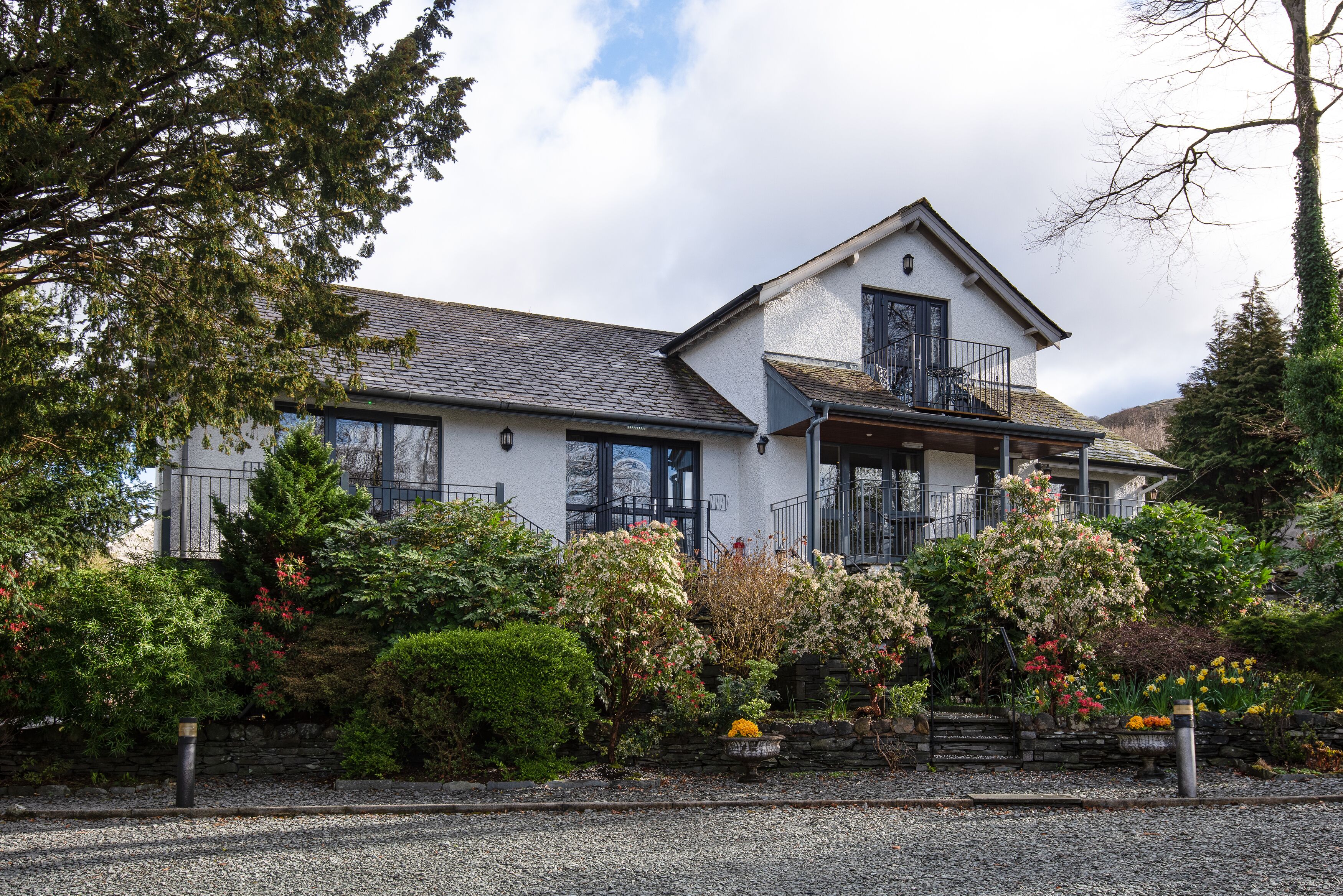 A two-story house with a balcony, surrounded by a well-maintained garden with various shrubs and flowering plants.