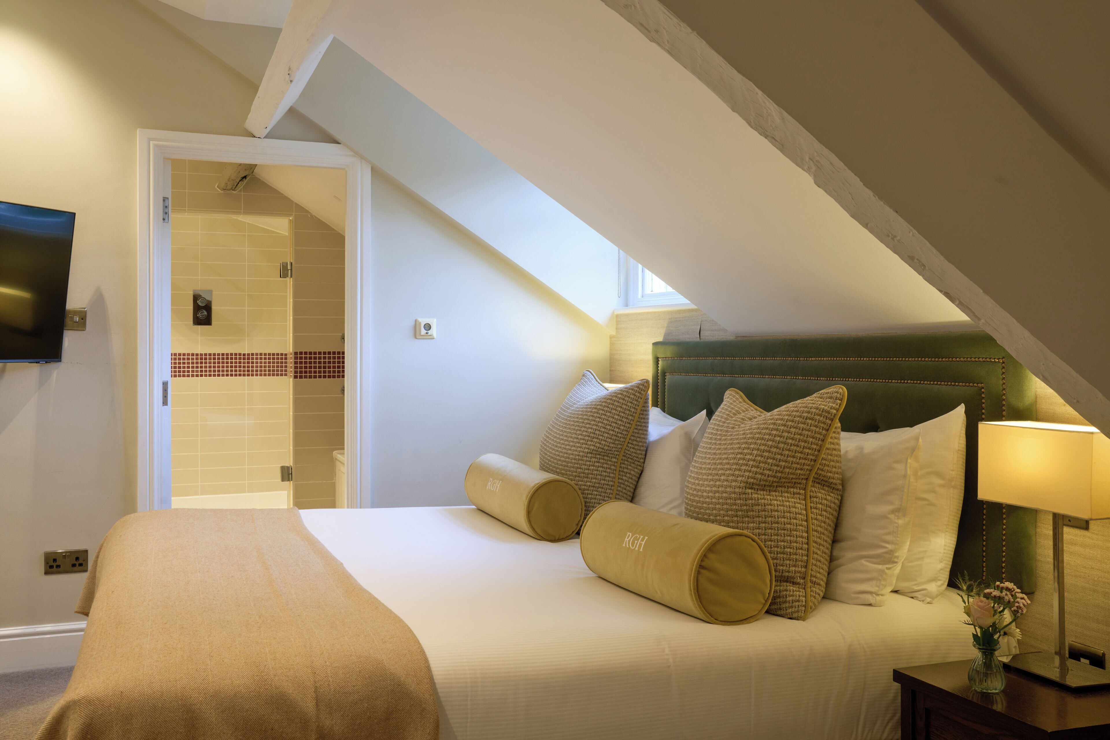 A hotel room with a bed, pillows, a nightstand, a lamp, and a television on the wall.