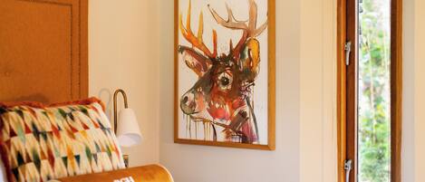 Een hotelkamer met een bed, een kleurrijk quilt, een schilderij van een hert en een raam met uitzicht op groen.