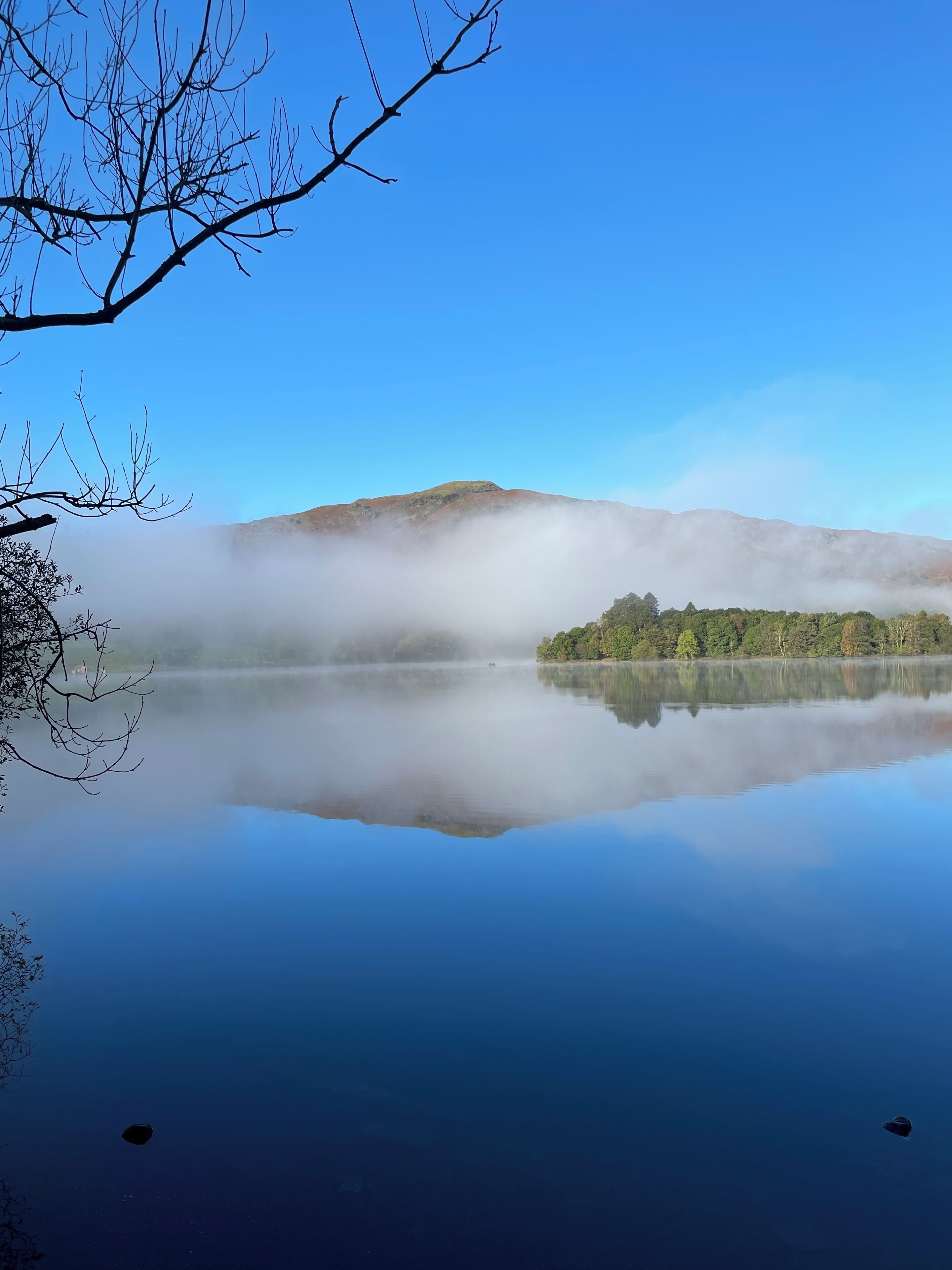 A calm lake with a clear reflection of the surrounding landscape, including a mountain and a small island with trees.