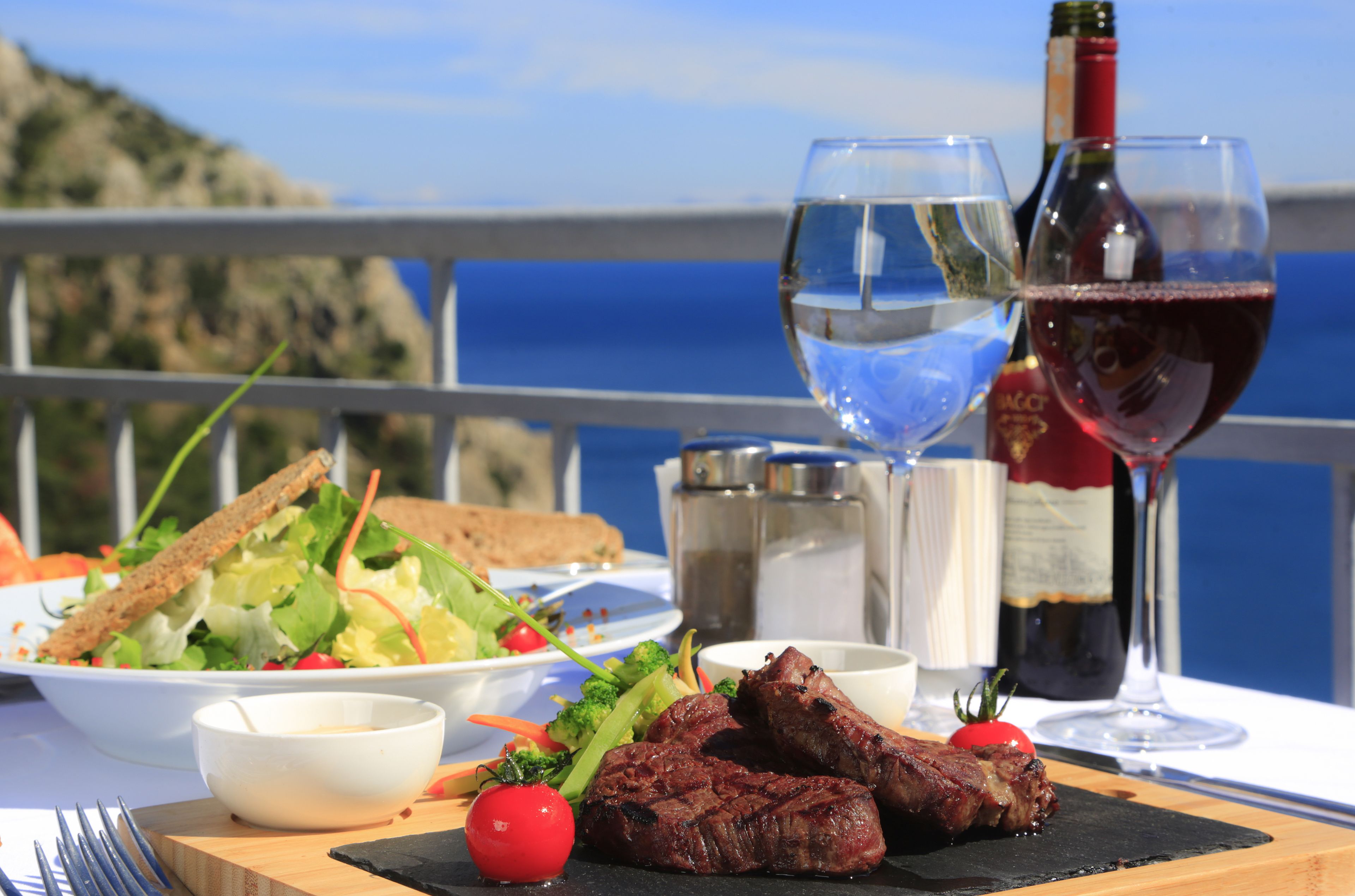 Une table dressée avec un repas composé de viande grillée, de salade et de pain, accompagné de verres à vin et d’une bouteille de vin.