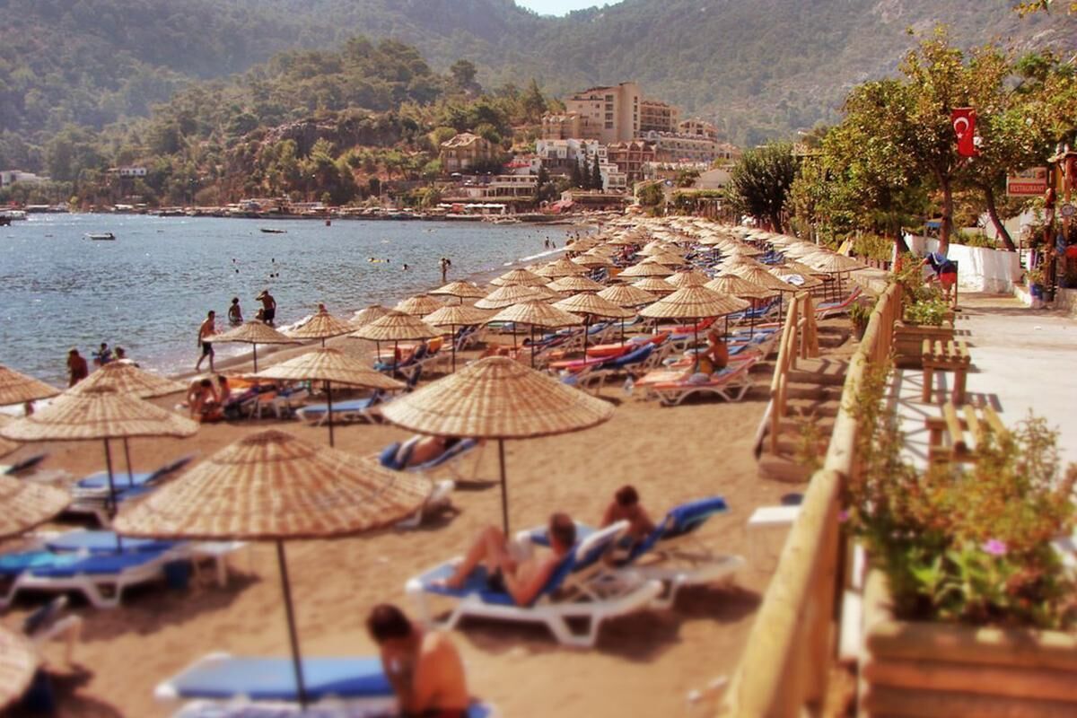 A beach with rows of sun loungers and thatched umbrellas, a clear view of the sea, and buildings in the background.