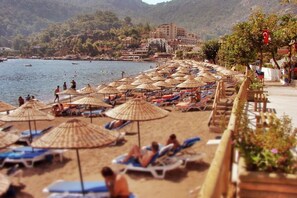 A beach with rows of sun loungers and thatched umbrellas, a clear view of the sea, and buildings in the background.