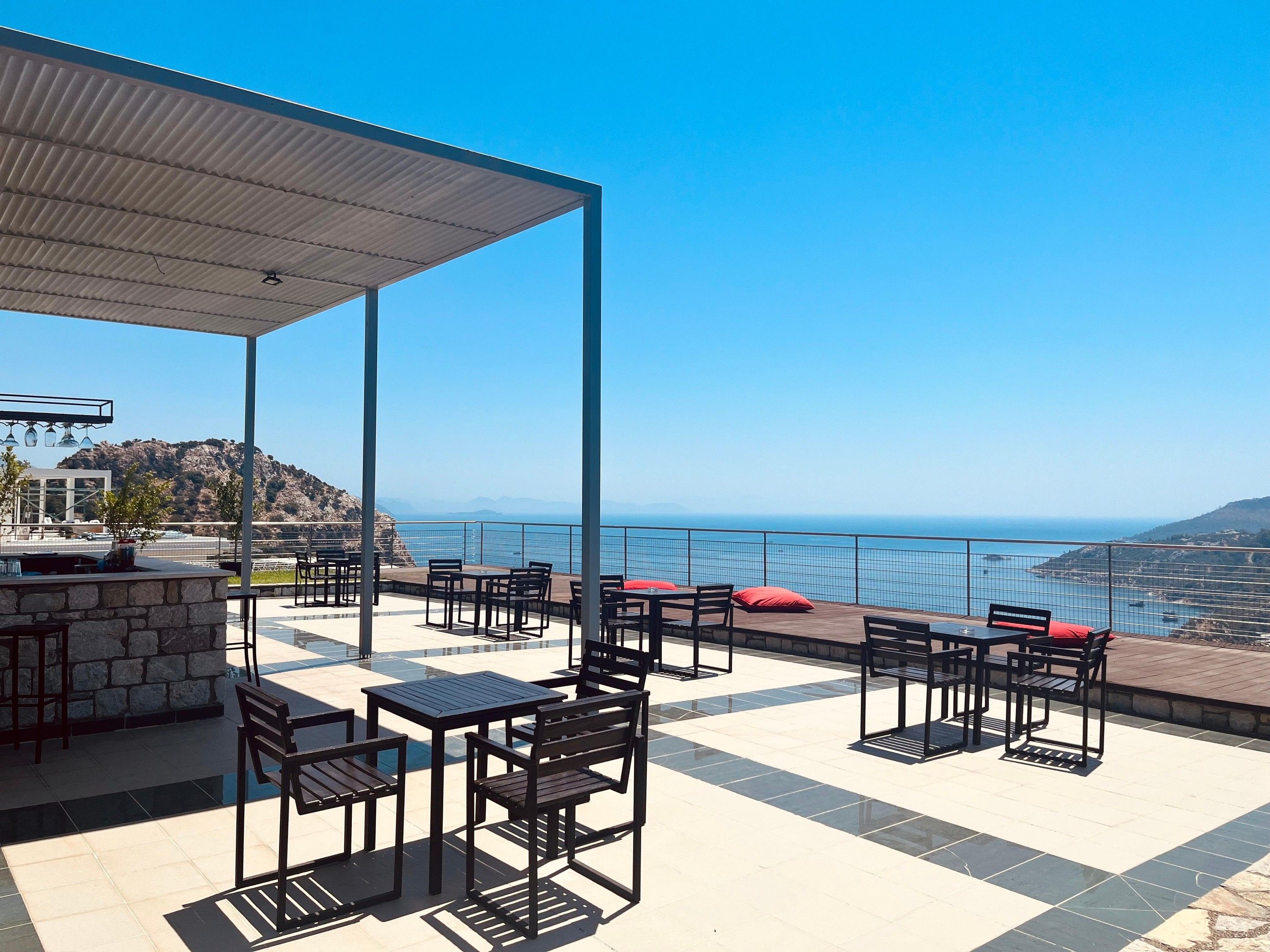 An outdoor patio with tables and chairs, a stone bar, and a view of the sea and mountains.