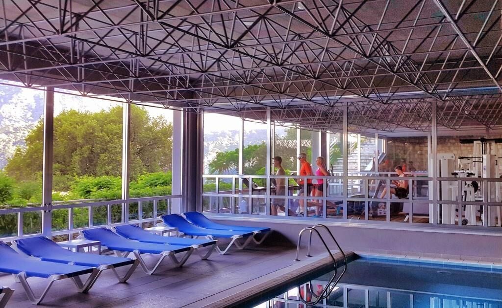 A covered swimming pool area with blue lounge chairs, large windows, and a view of greenery outside.