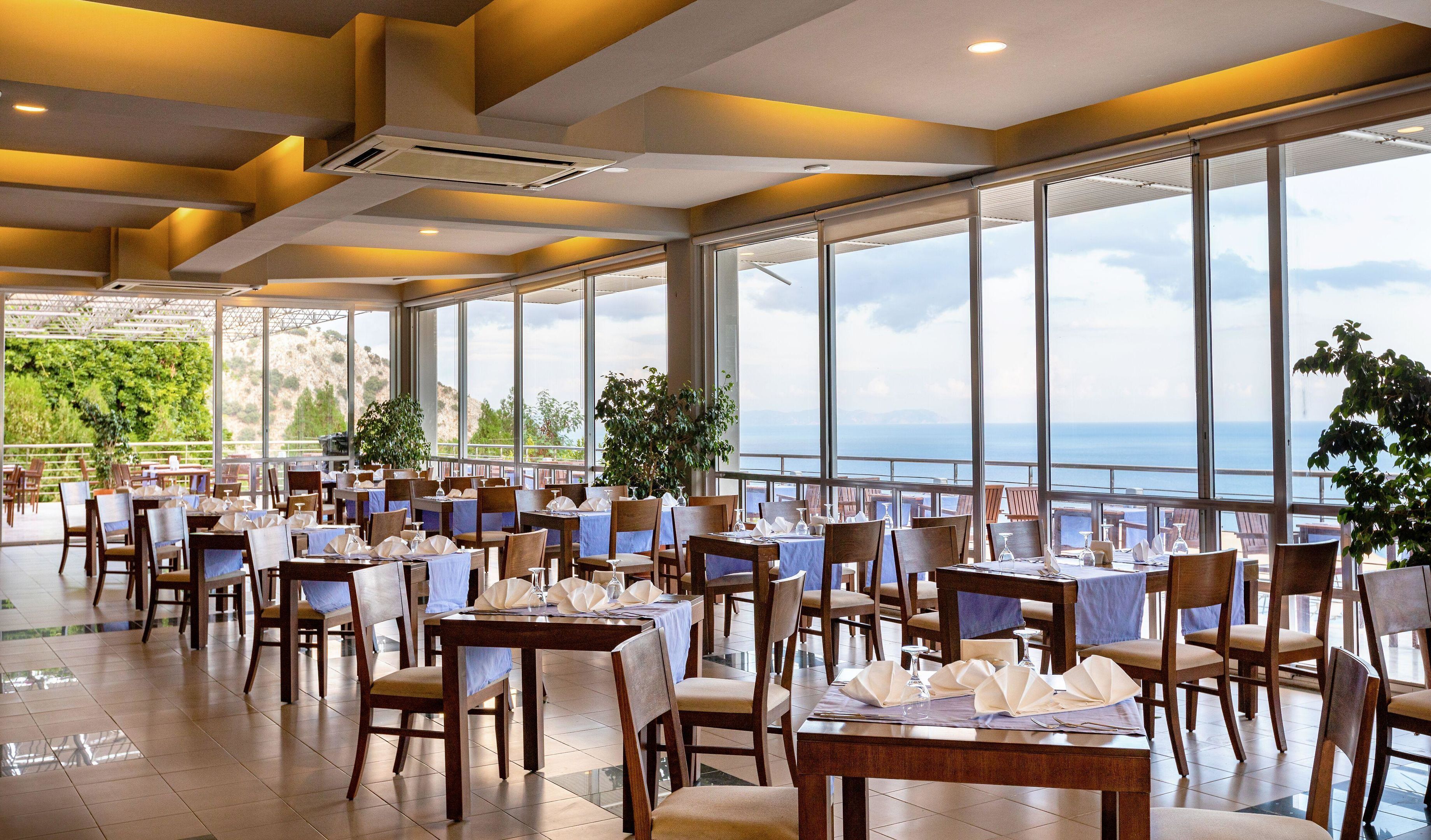 A restaurant with tables set for dining, large windows offering a view of the sea, and potted plants.
