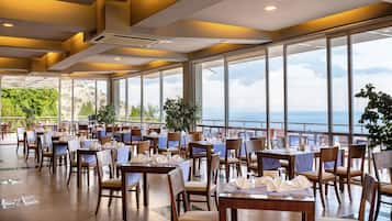 Un restaurante con mesas dispuestas para comer, amplias ventanas que dan al mar y plantas en macetas.
