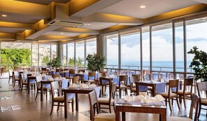 Un restaurant avec des tables dressées pour le service, de grandes fenêtres donnant sur la mer et des plantes en pot.