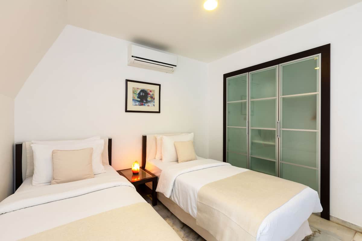 Habitación de hotel con dos camas, una mesita de noche, aire acondicionado, un cuadro en la pared y un armario con puerta corrediza de vidrio.