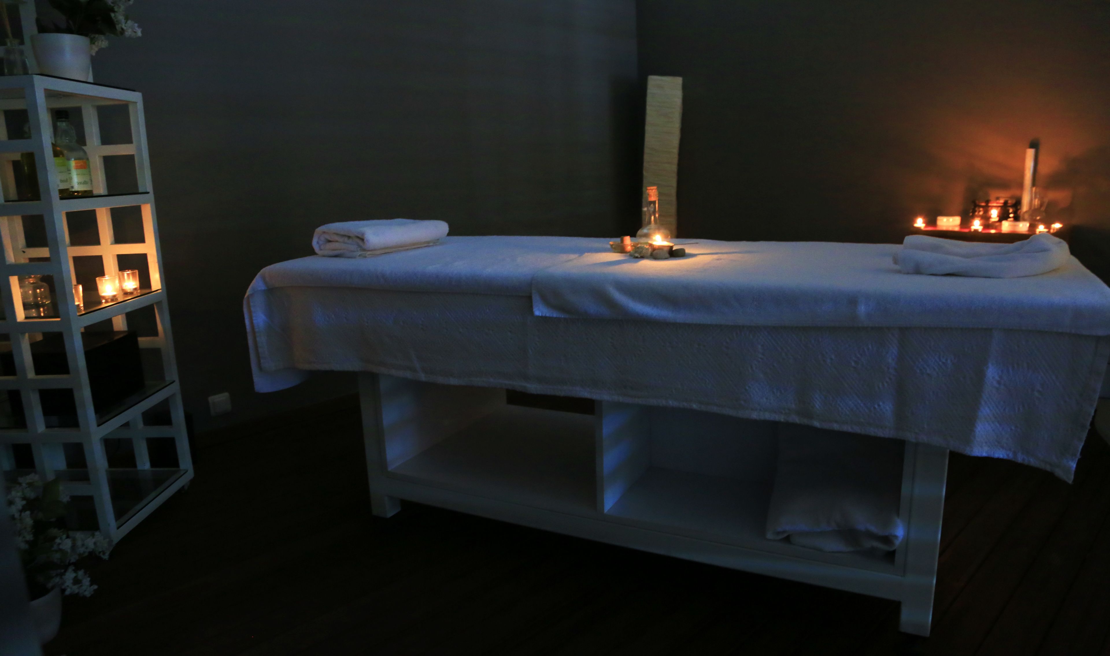 Une salle de soins avec une table de massage, des serviettes, des bougies et une étagère où sont disposées des bouteilles et des fleurs.