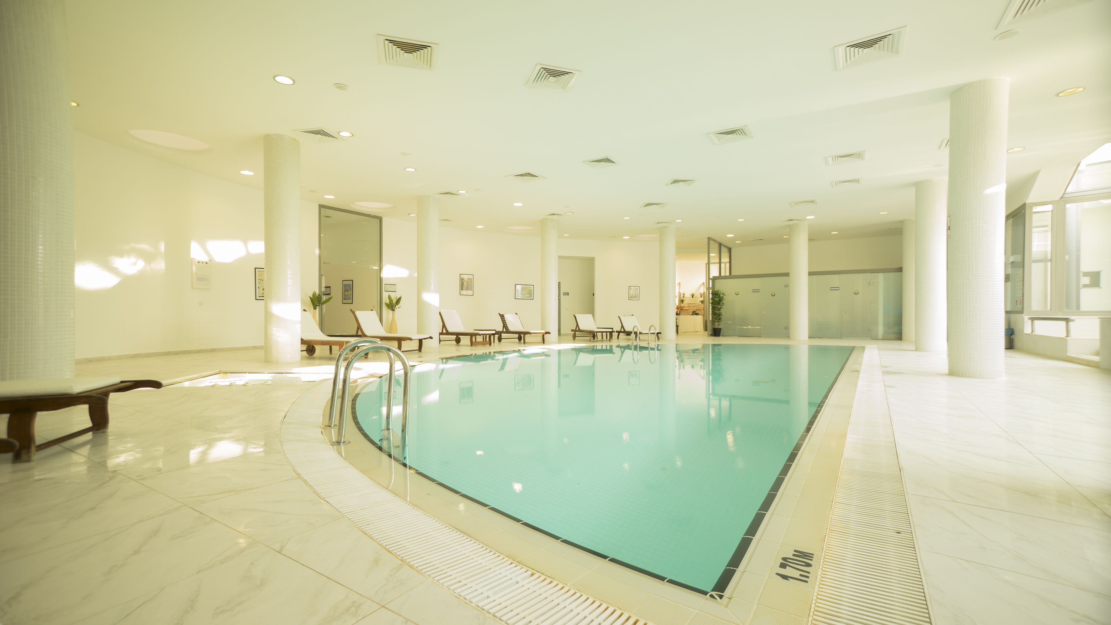 A large indoor swimming pool with lounge chairs and a bench, surrounded by white columns and a tiled floor.