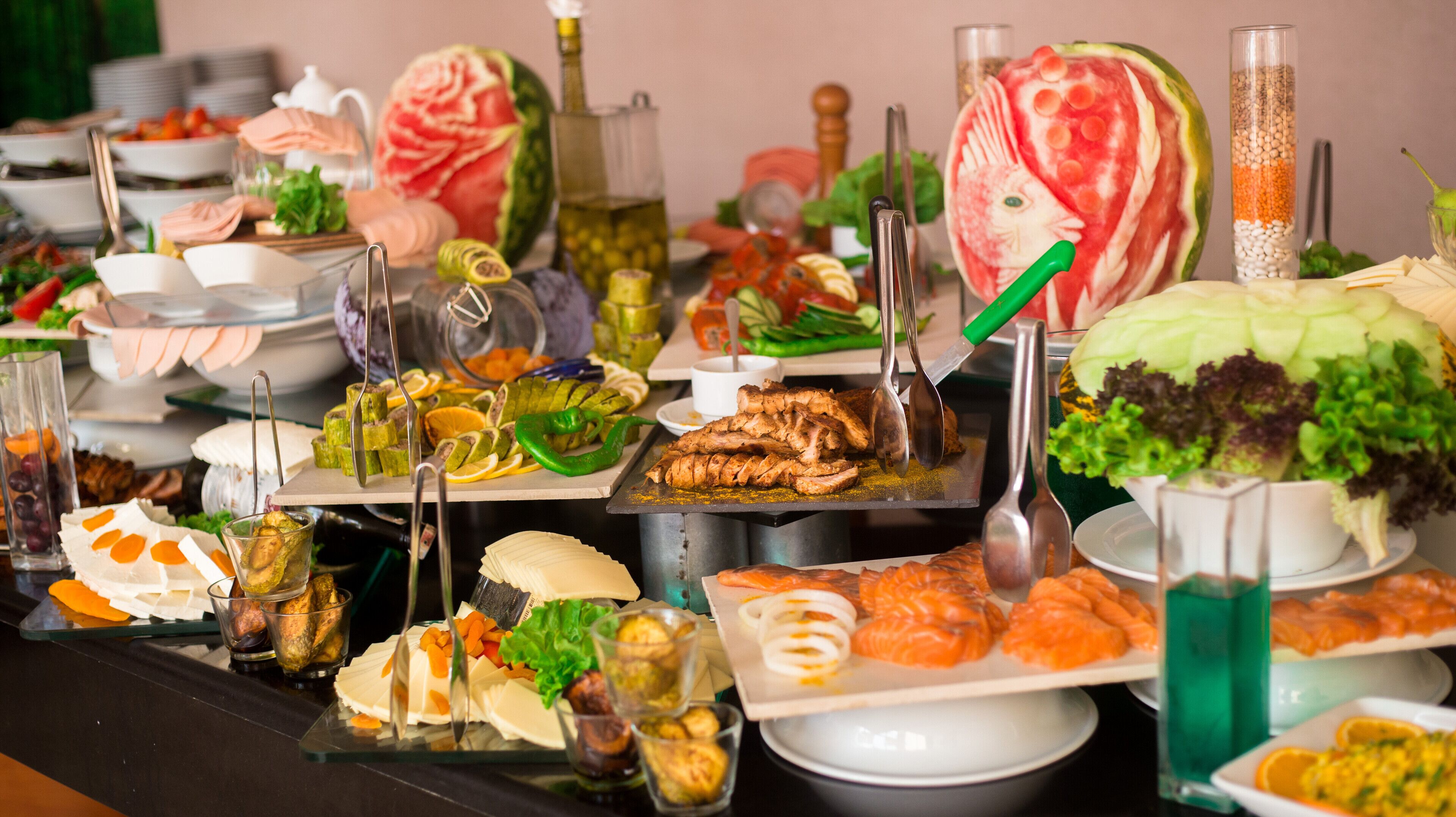 Una mesa de buffet con diversos platos, entre ellos, carnes cortadas, verduras y una sandía tallada como pez.