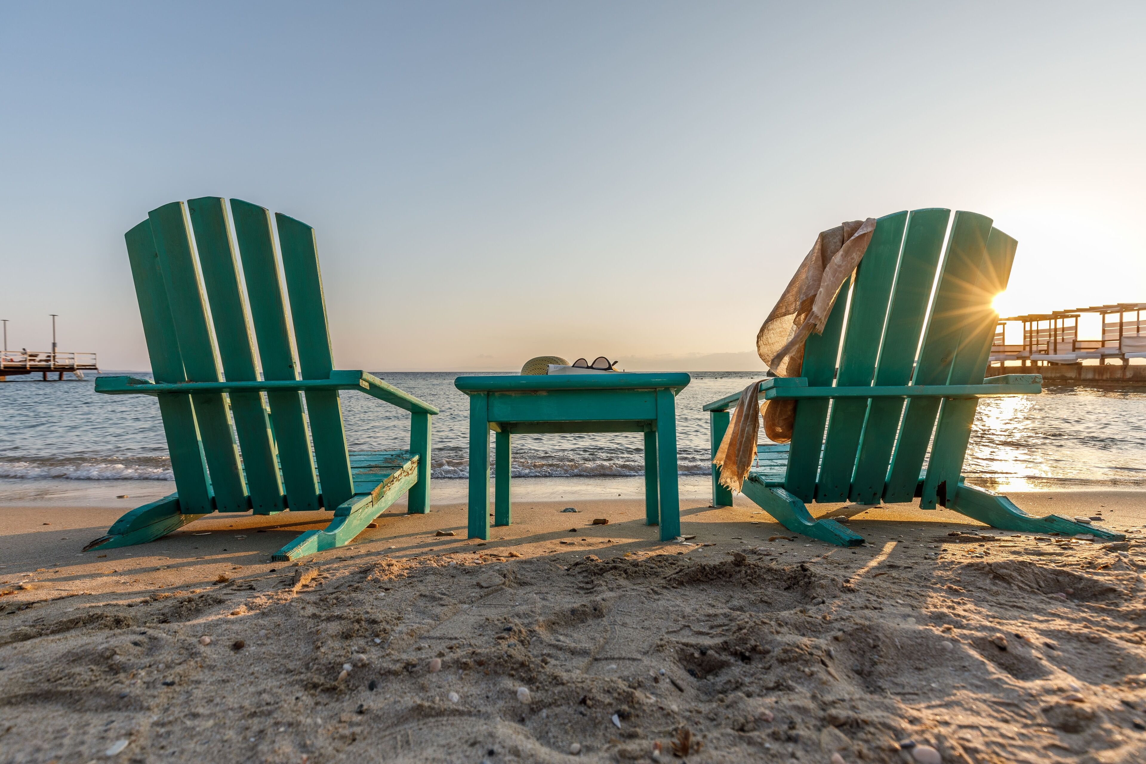 Dos sillas Adirondack verdes y una mesa pequeña sobre una playa arenosa, con el océano al fondo.