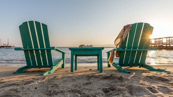 Dos sillas Adirondack verdes y una mesa pequeña sobre una playa arenosa, con el océano al fondo.
