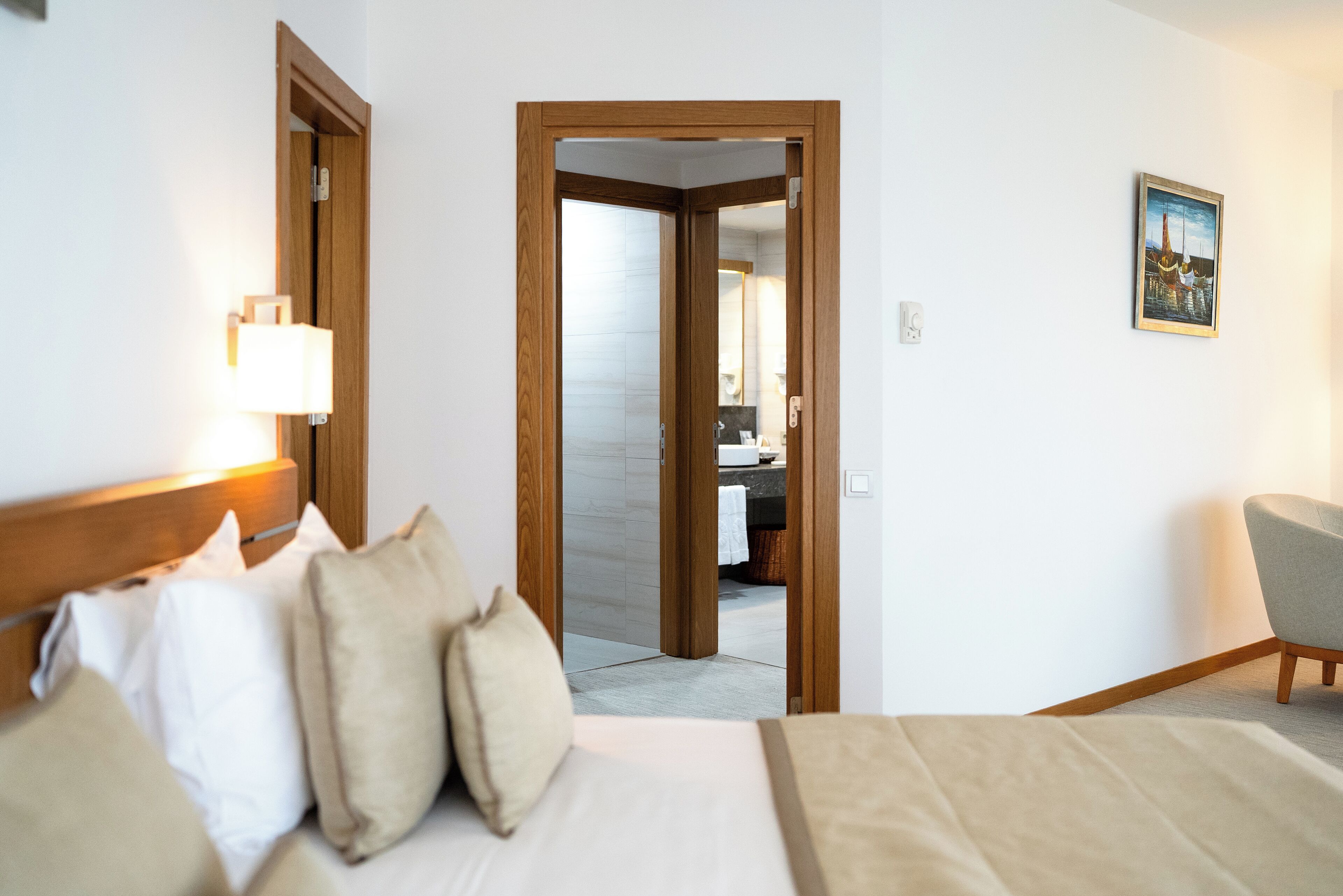 A hotel room with a bed, a nightstand, a lamp, a chair, and a bathroom visible through an open door.