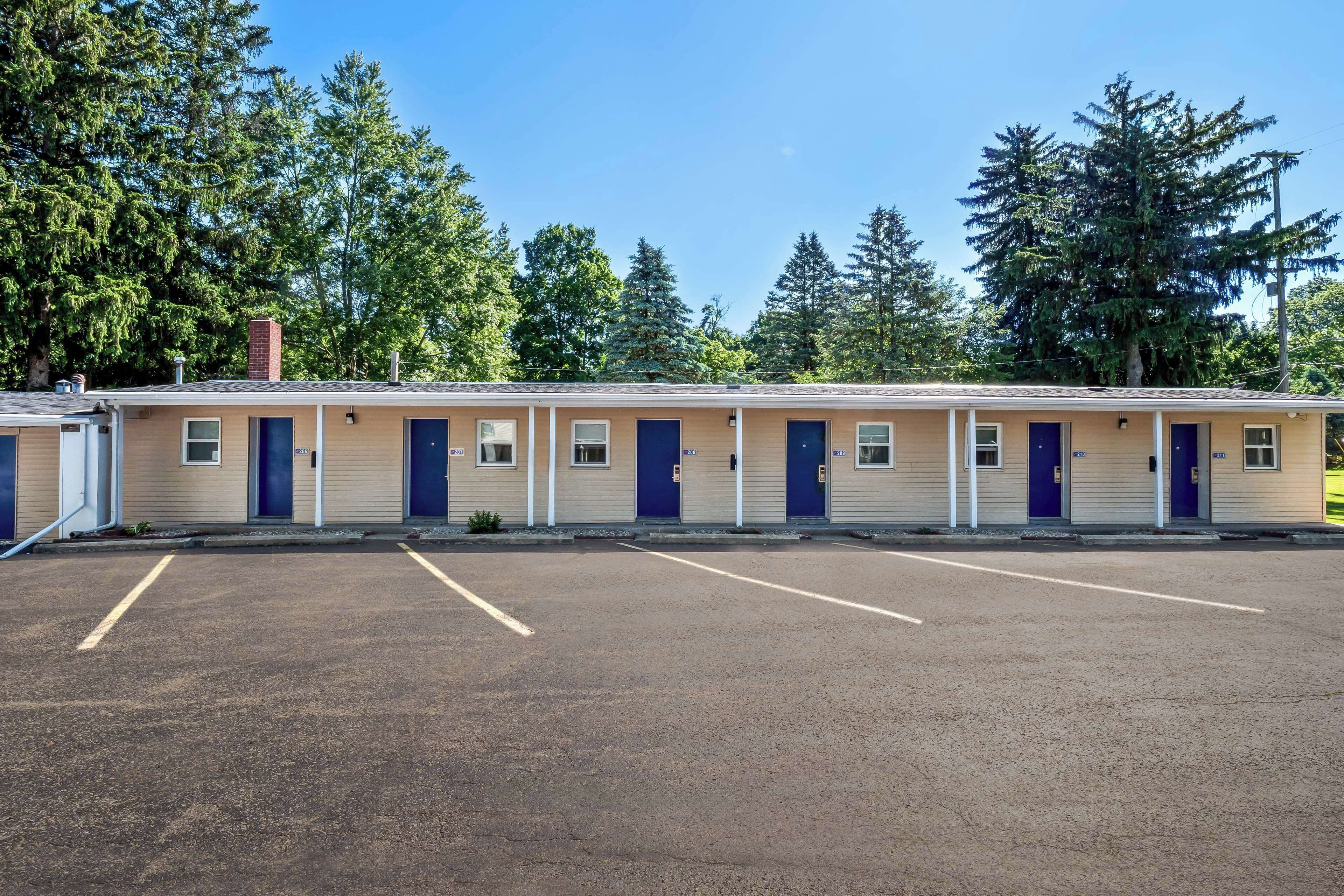 A row of motel rooms with individual doors, a parking lot, and trees in the background.