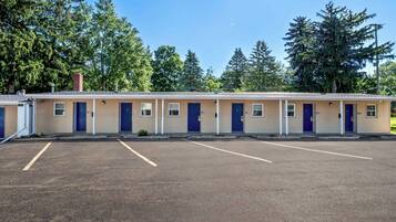 A row of motel rooms with individual doors, a parking lot, and trees in the background.