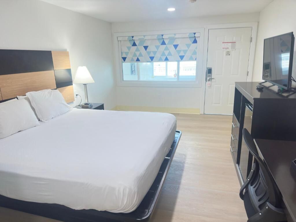 A hotel room with a bed, a desk with a computer, a TV, a window with blinds, and a door.