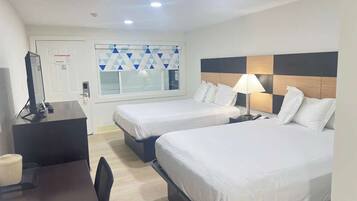 A hotel room with two beds, a desk, a chair, a lamp, and a window with geometric curtains.