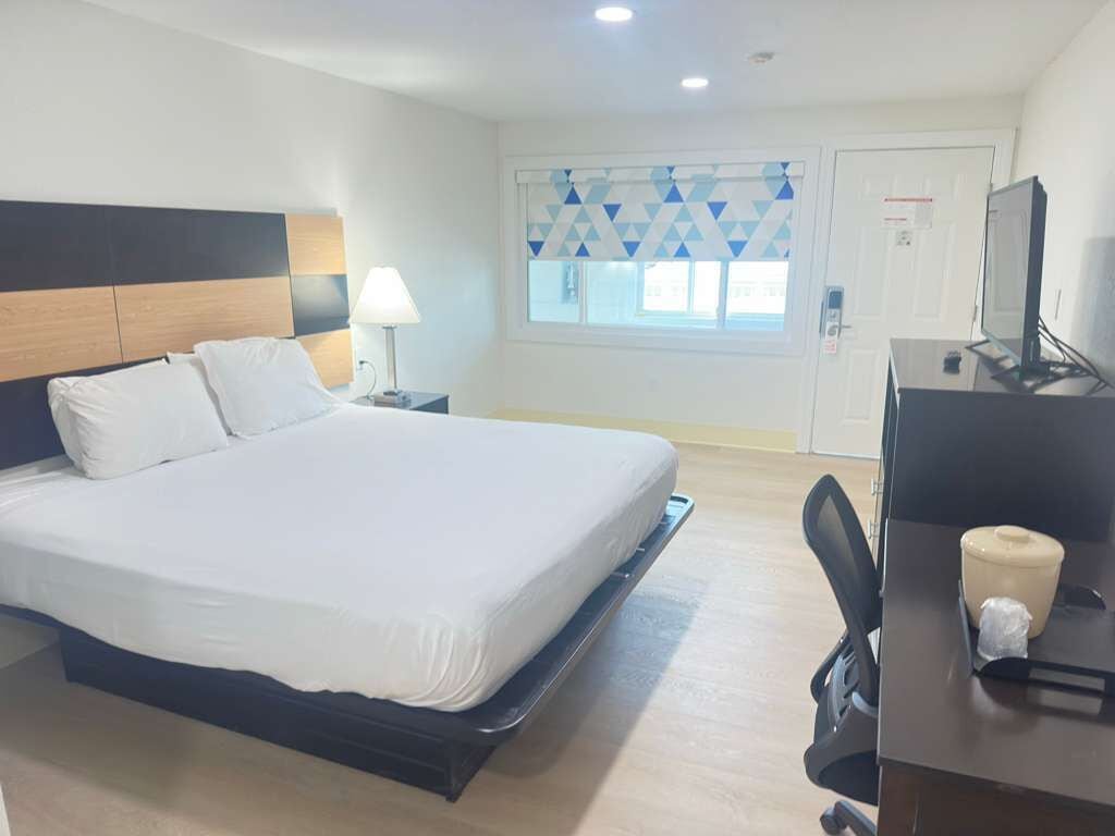 Una habitació d'hotel amb un llit, una taula de nit, una làmpada, una finestra amb persianes geomètriques, un escriptori amb un ordinador i una televisió.