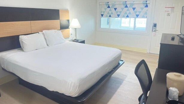 A hotel room with a bed, bedside table, lamp, window with blinds, and a desk with a chair. - Bourne Bridge Inn  (Buzzards Bay)