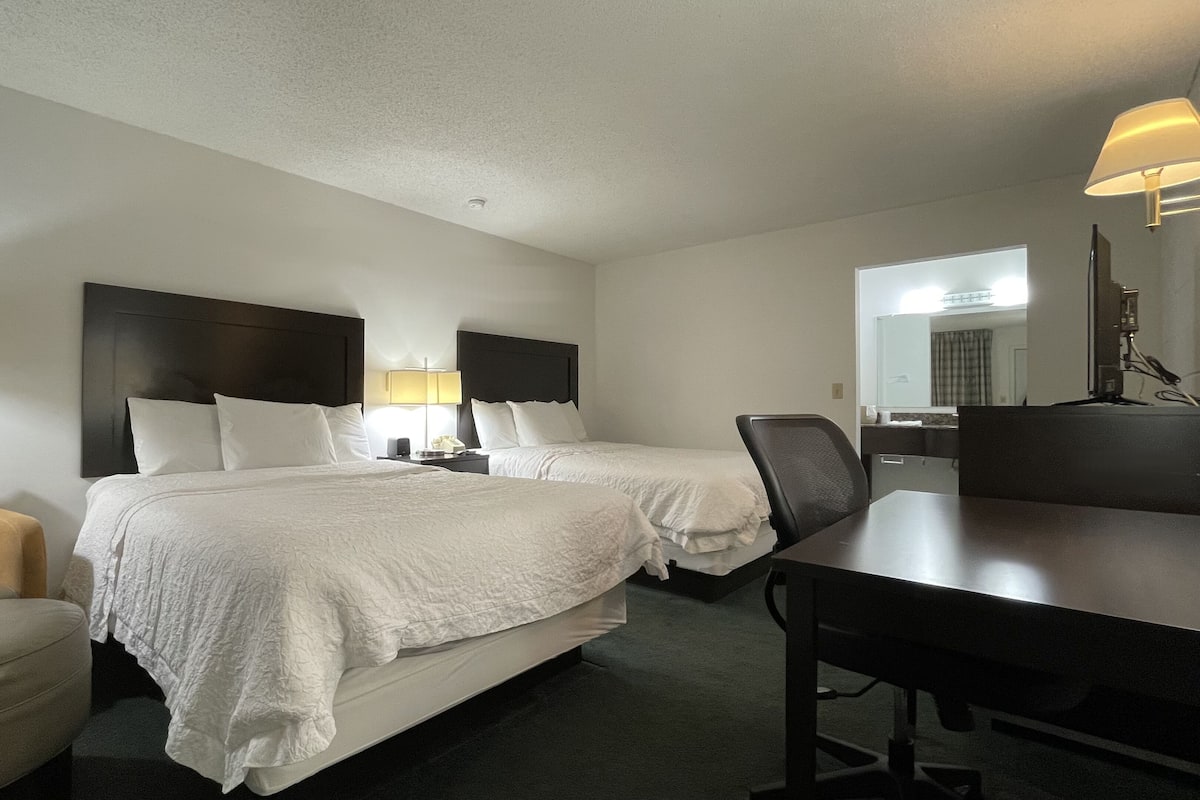 A hotel room with two beds, a desk, a chair, a sofa, and a lamp.