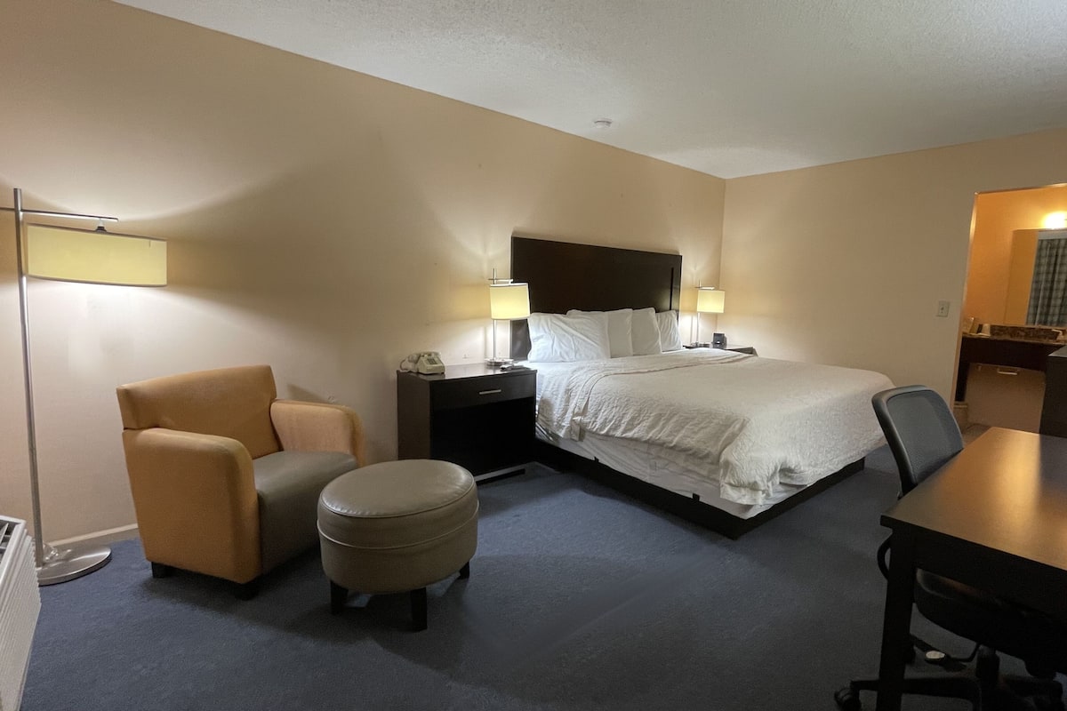 A hotel room with a bed, a desk, a chair, a small ottoman, a nightstand, and a lamp.