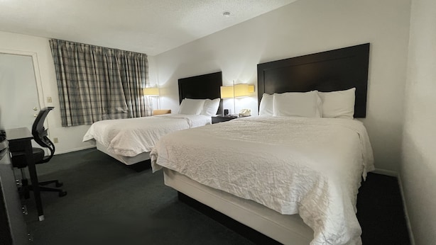 A hotel room with two beds, a desk, a chair, and a window with plaid curtains. - Slumber Inn Of Harrisonville (Harrisonville)