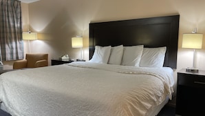 A hotel room with a large bed, two bedside tables, a chair, and a window with curtains. - Slumber Inn Of Harrisonville (Harrisonville)