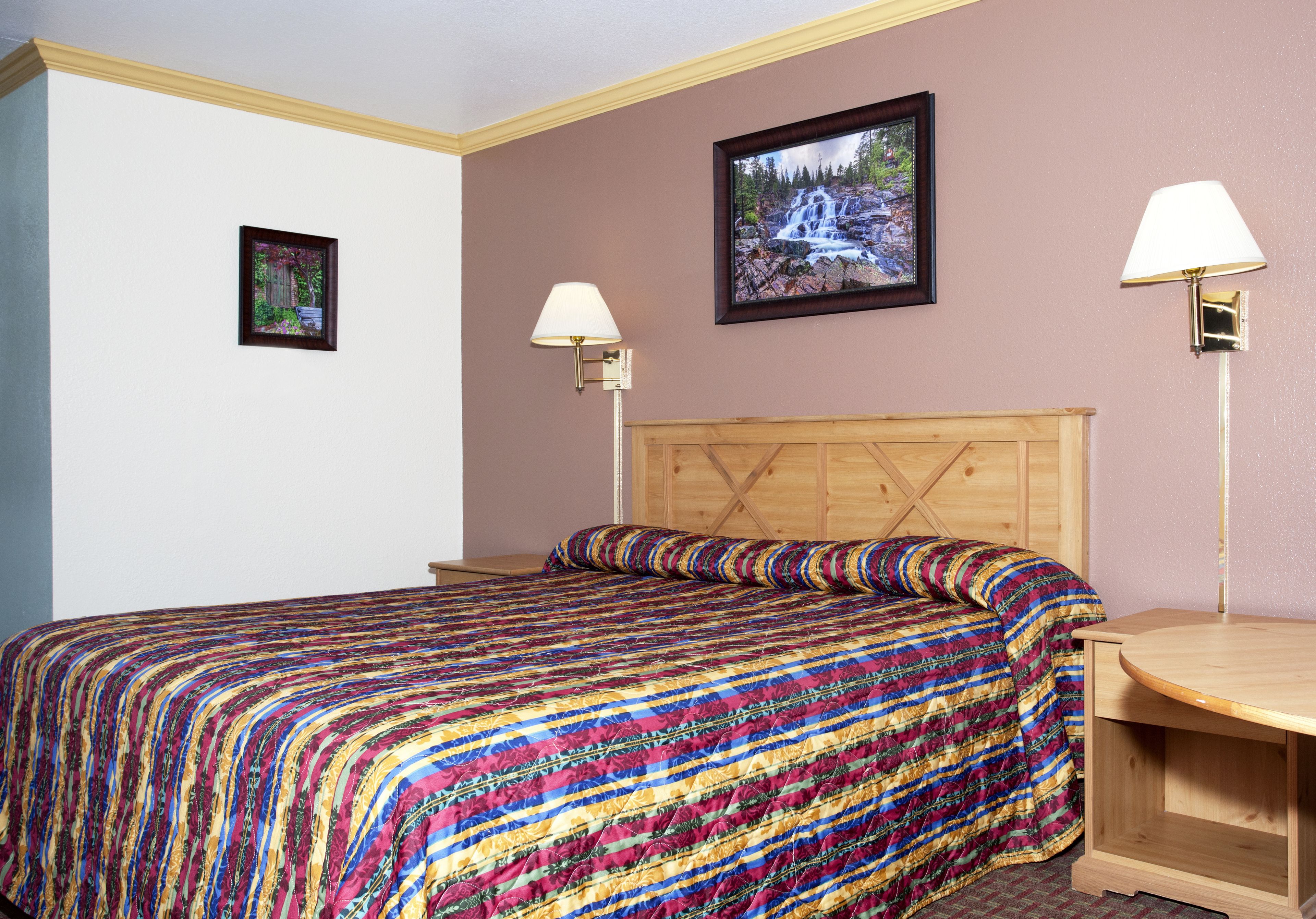 A hotel room with a wooden headboard, a colorful striped bedspread, two bedside lamps, and a framed picture of a waterfall on the wall.