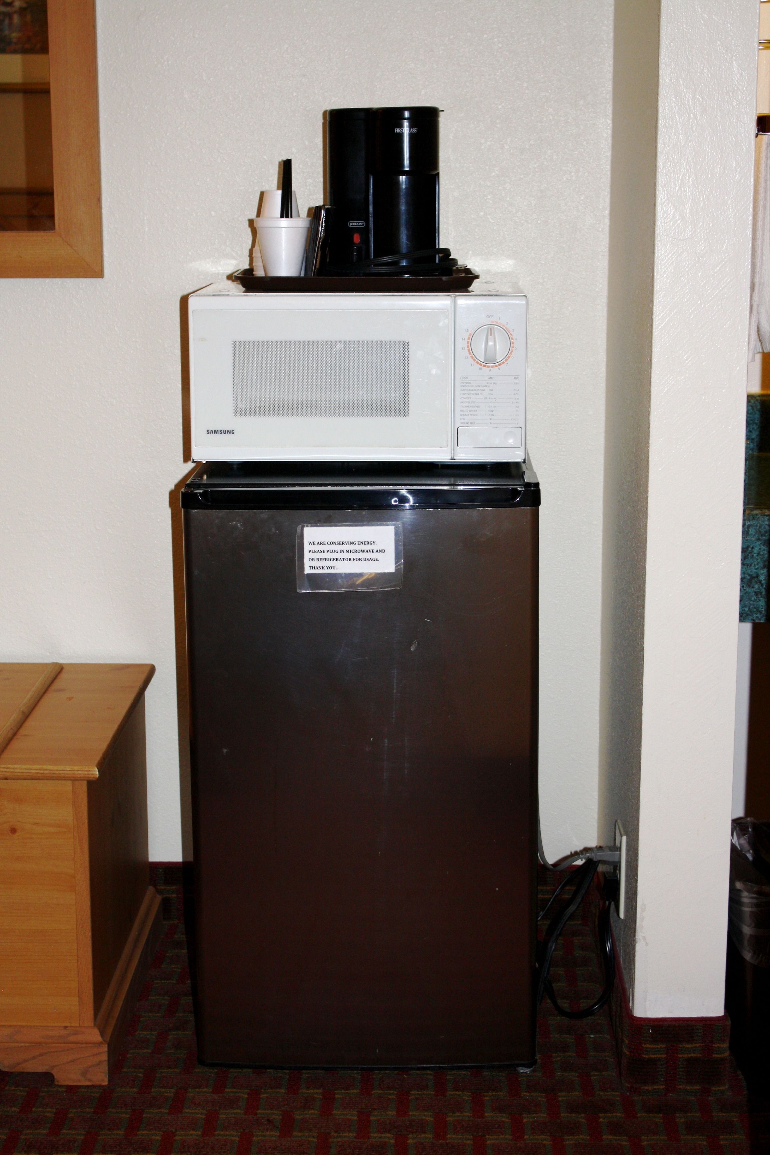 Un refrigerador compacto con un microondas y una cafetera encima, una etiqueta de advertencia en la puerta y un armario de madera a la izquierda.