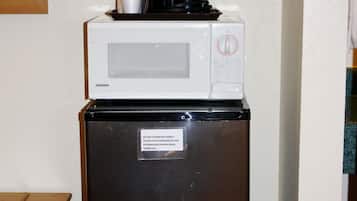 Un refrigerador compacto con un microondas y una cafetera encima, una etiqueta de advertencia en la puerta y un armario de madera a la izquierda.