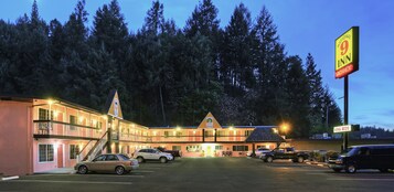 Un motel con un edificio de dos pisos, un estacionamiento y un paisaje boscoso al fondo.