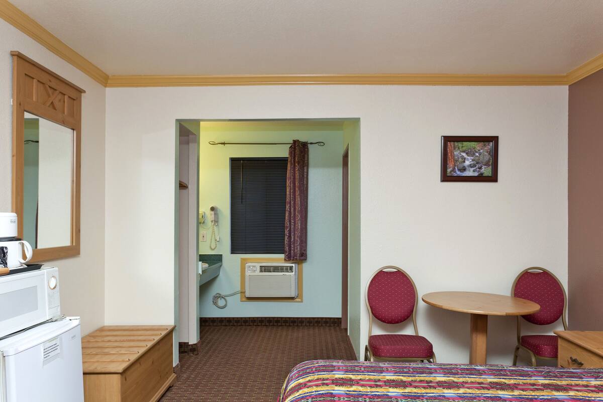 A hotel room with a bed, a small table with two chairs, a microwave, a refrigerator, a bathroom visible through an open door, and a picture on the wall.