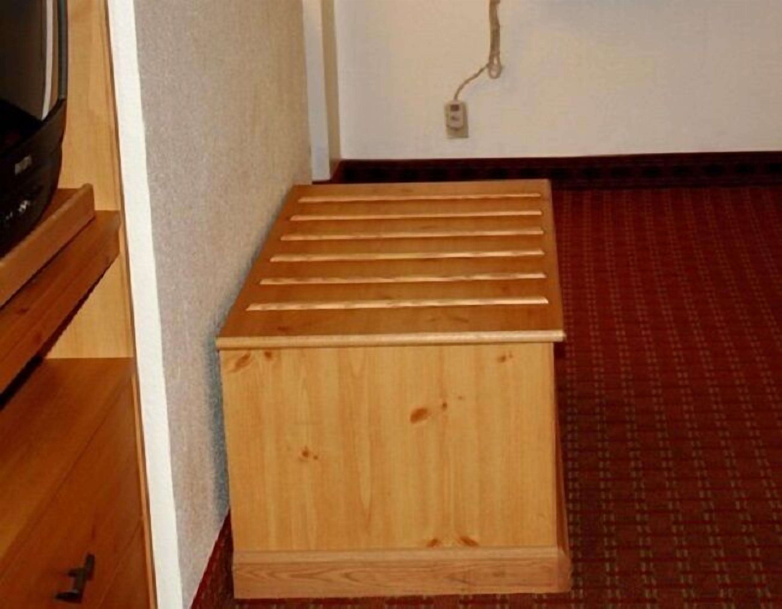 Un petit meuble en bois, doté d’un dessus à lattes, placé dans une pièce dont le sol est recouvert d’un tapis à motifs et dont un des murs est blanc.