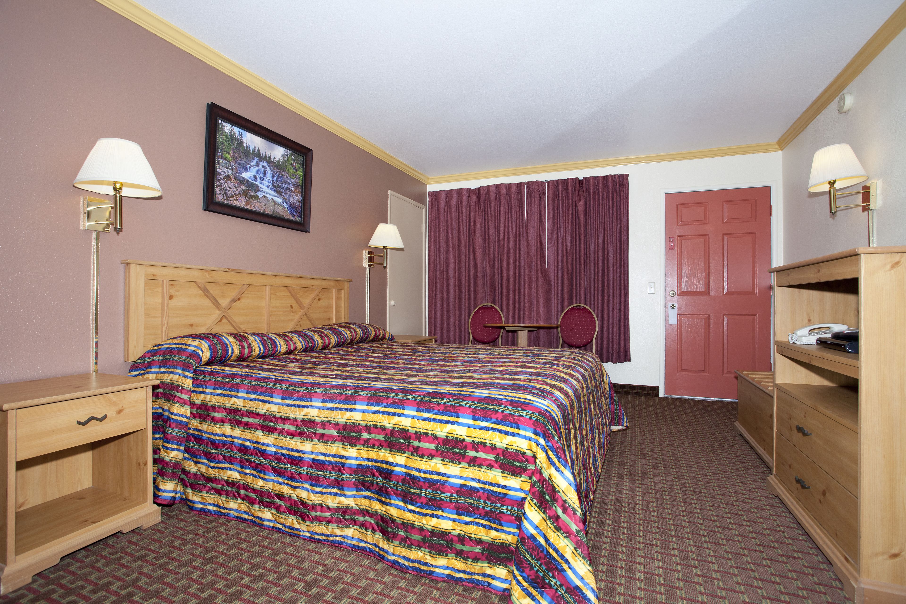 Habitación de hotel con una cama de madera, dos mesitas de noche, un escritorio con una silla, una puerta roja y un cuadro enmarcado en la pared.