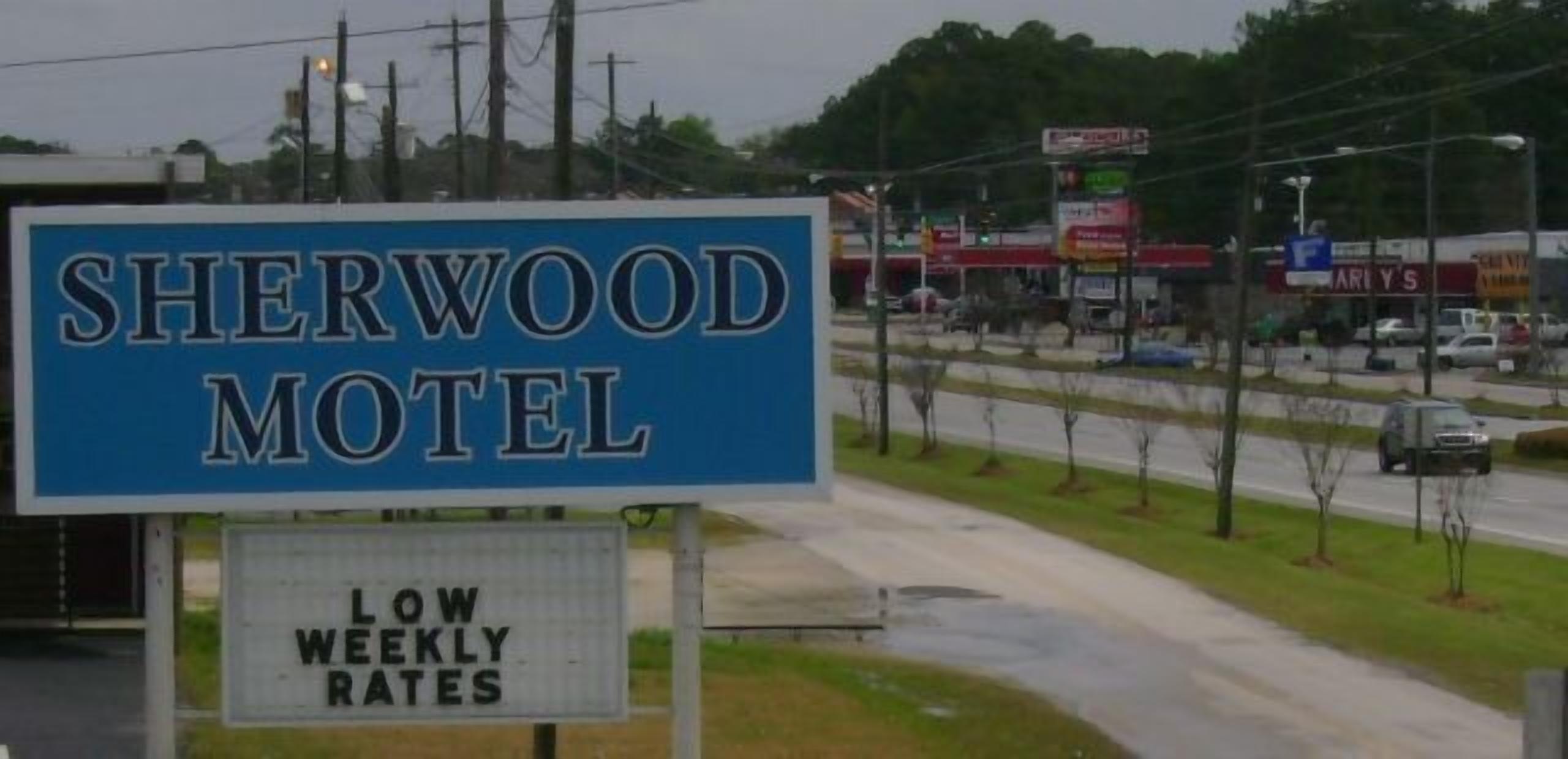 Letrero de motel que dice "SHERWOOD MOTEL" y un cartel debajo que indica "TARIFA SEMANAL ECONÓMICA".