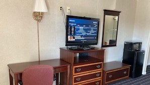 Habitación de hotel con un escritorio de madera, un televisor mostrando una guía de canales, una silla de oficina roja, un espejo y un refrigerador negro.