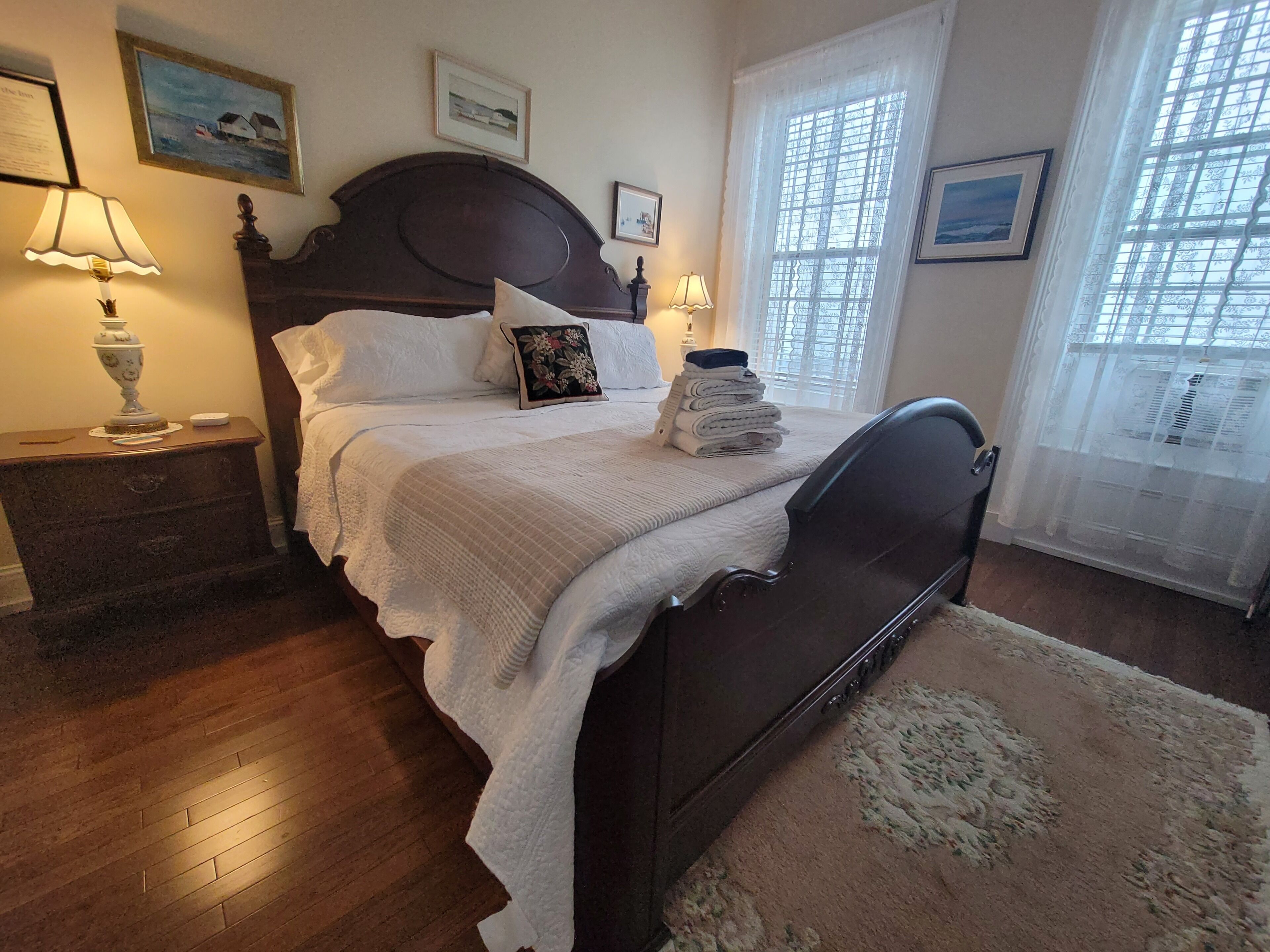 A neatly made bed with white linens, a dark wooden headboard, and a floral pillow. There are two bedside lamps, a framed picture above the bed, and a window with a sheer curtain.