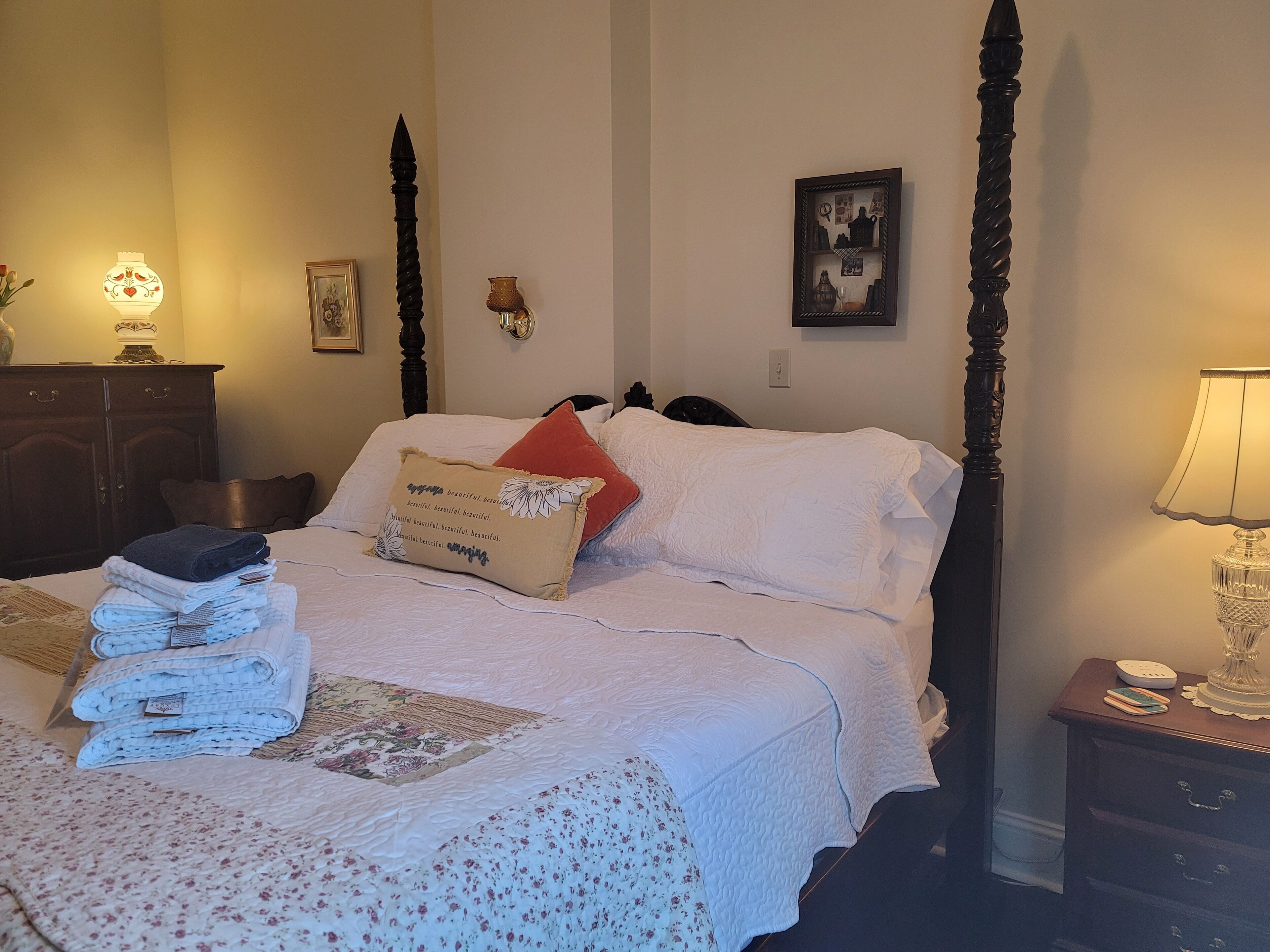 A neatly made bed with a floral-patterned bedspread, a stack of folded towels, and a decorative pillow with a floral design.