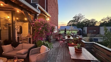 Uma área de pátio externa com móveis de madeira, plantas em vasos e vista para um edifício ao fundo.