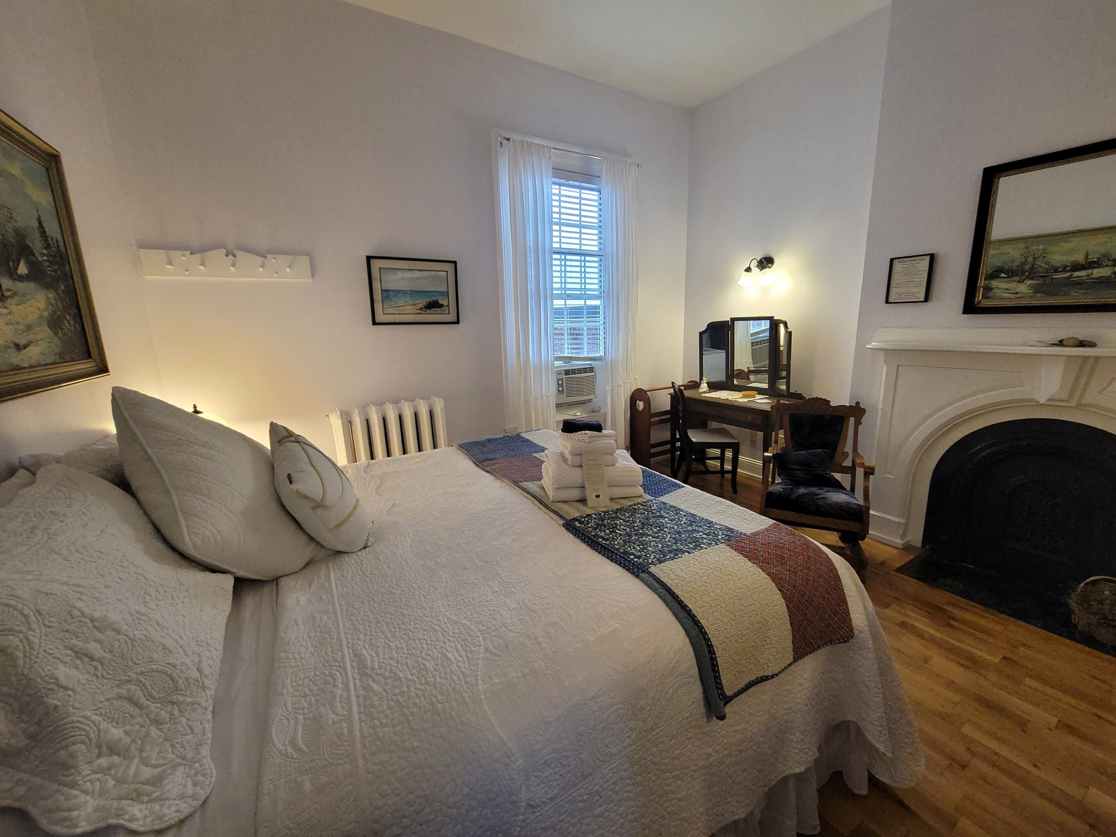 A neatly arranged bedroom with a bed, a desk, a fireplace, and a window with blinds.
