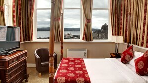 A hotel room with a large bed, a television, a chair, and a view of a cityscape through curtains. - The Argyll Hotel (Dunoon)