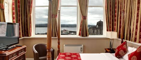 Habitación de hotel con una cama grande, televisión, una silla y vistas a un paisaje urbano a través de cortinas.